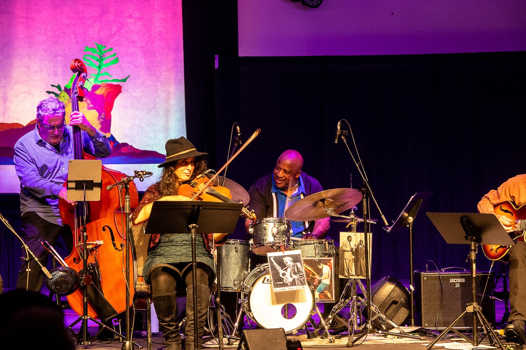 A jazz band performs energetically on stage. The bassist, violinist, drummer, and guitarist are illuminated by colorful lights, conveying a lively, joyful atmosphere.