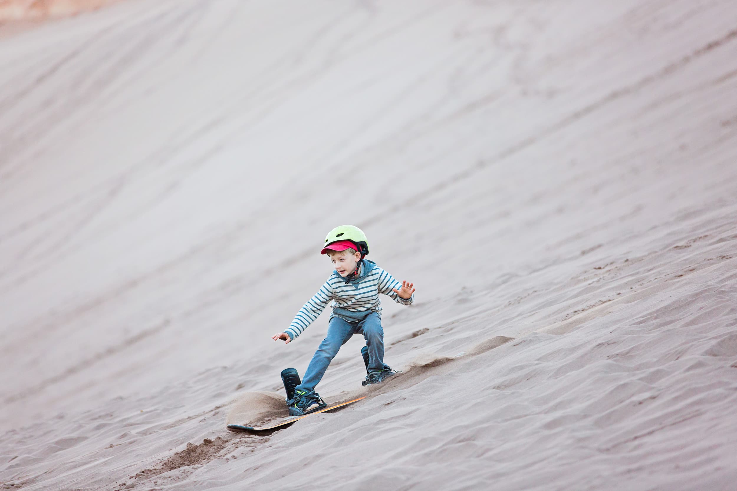A young child wearing a helmet dirt boarding down a sandy slope, with a determined expression. The desert background is vast and smooth, conveying adventure.