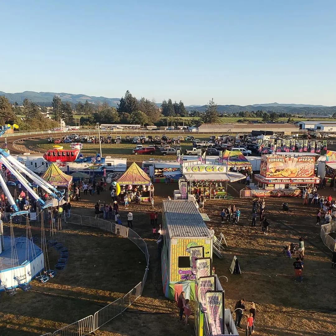 Aerial view of a vibrant fairground with colorful tents and bustling crowds. Rides and food stalls are scattered, set against a backdrop of trees and mountains.