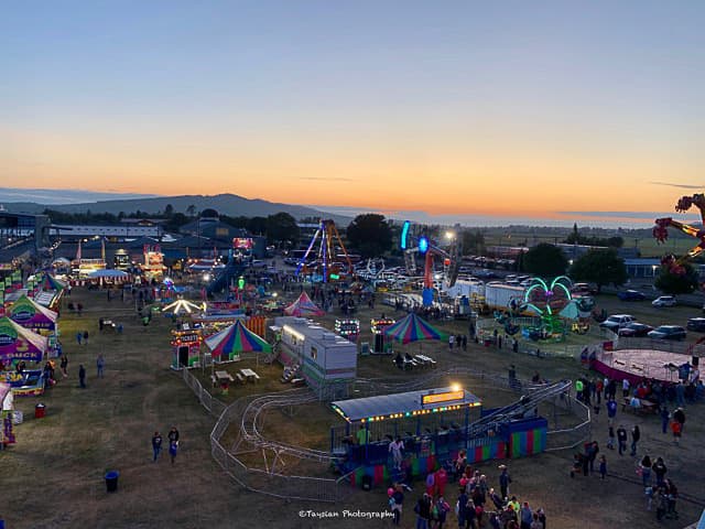 Aerial view of a vibrant fairground at sunset. Colorful tents, rides, and lively crowds are seen. The sky is orange and blue, evoking a festive mood.