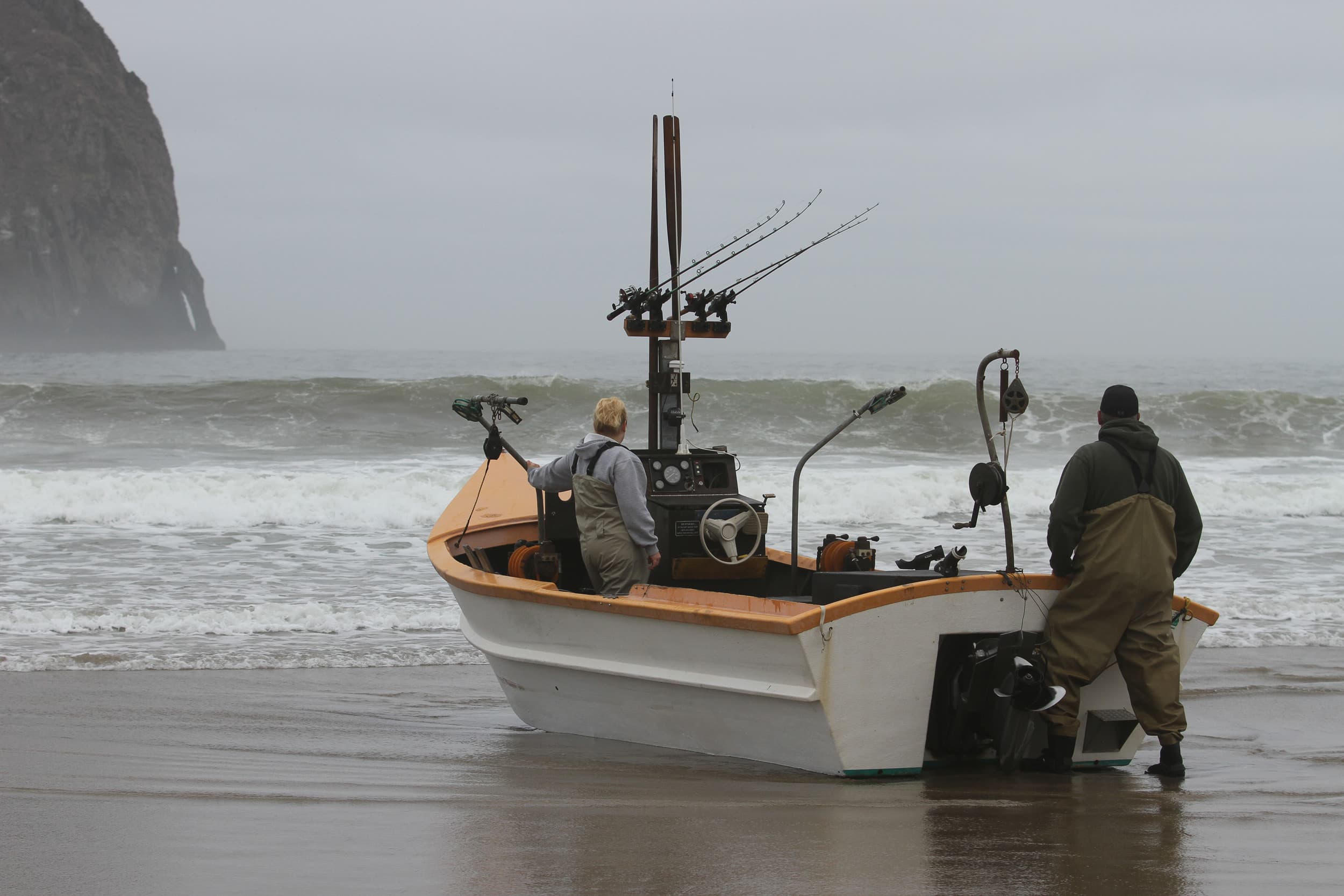 Two people stand in a small fishing boat on a gray, overcast beach. Fishing rods are visible, and waves crash gently in the background.