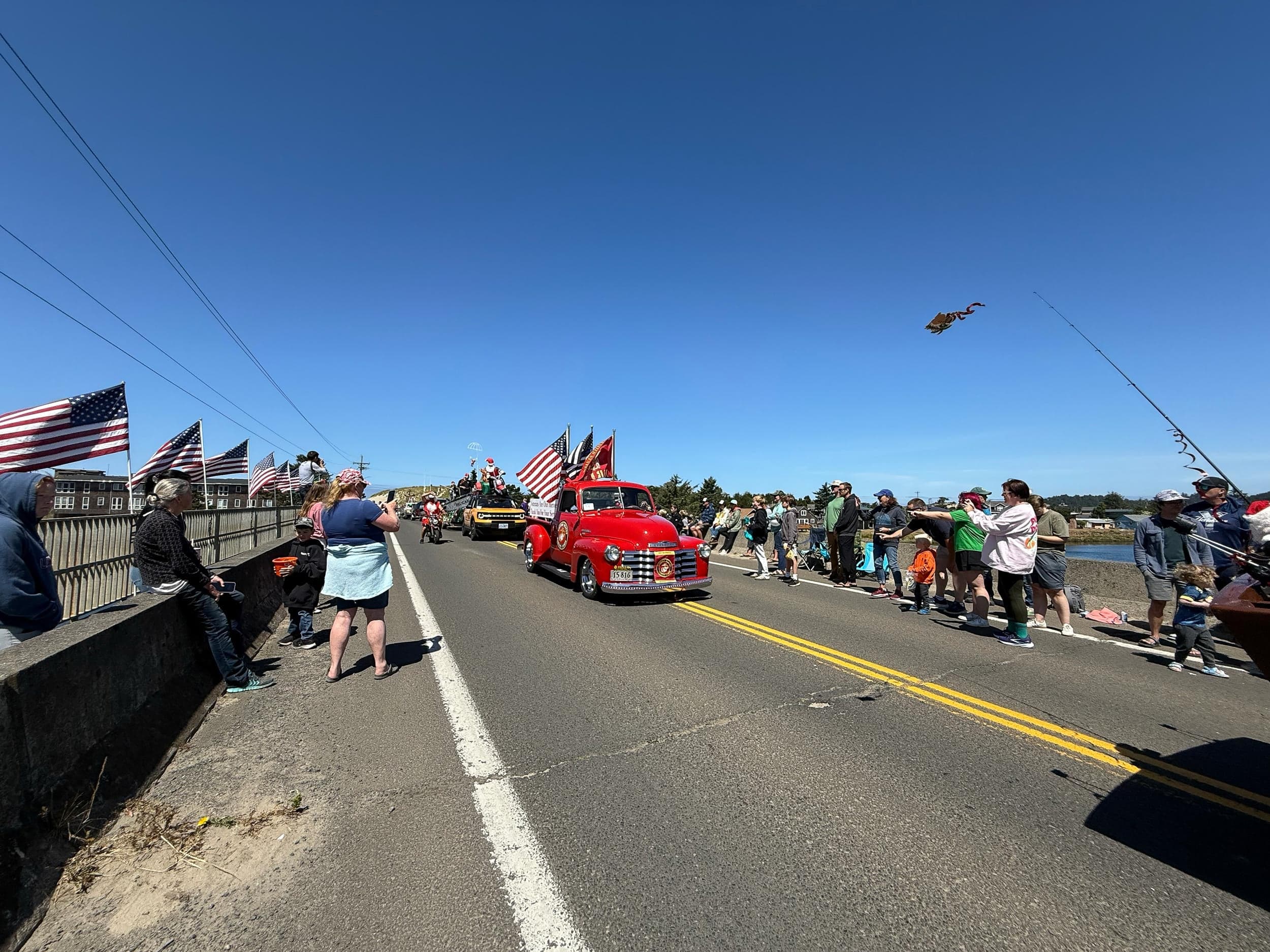 A vibrant parade on a sunny day features a classic red truck with American flags. Spectators line the street, creating a festive, patriotic atmosphere.