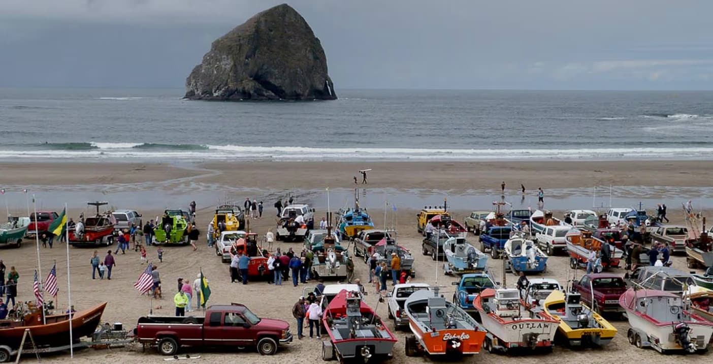 A busy beach scene with colorful cars and boats parked on the sand, people gathered around, and a large rock formation in the ocean under a cloudy sky.
