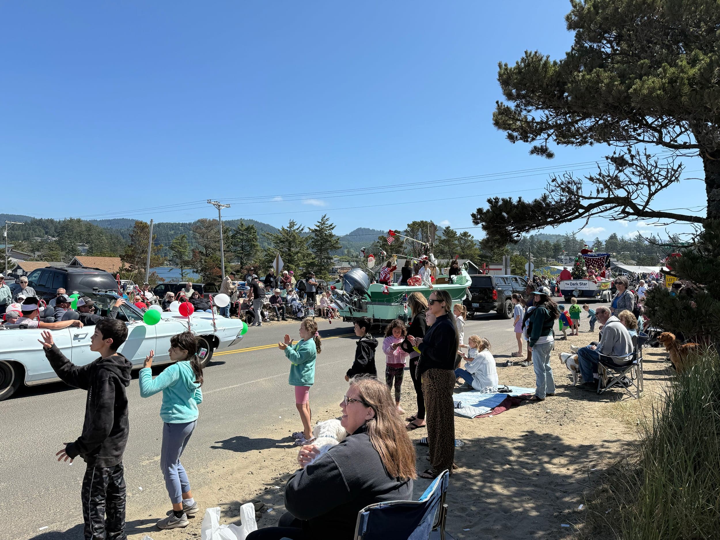 A lively parade on a sunny day with classic cars adorned with balloons, people waving, children watching, and spectators seated along the road.