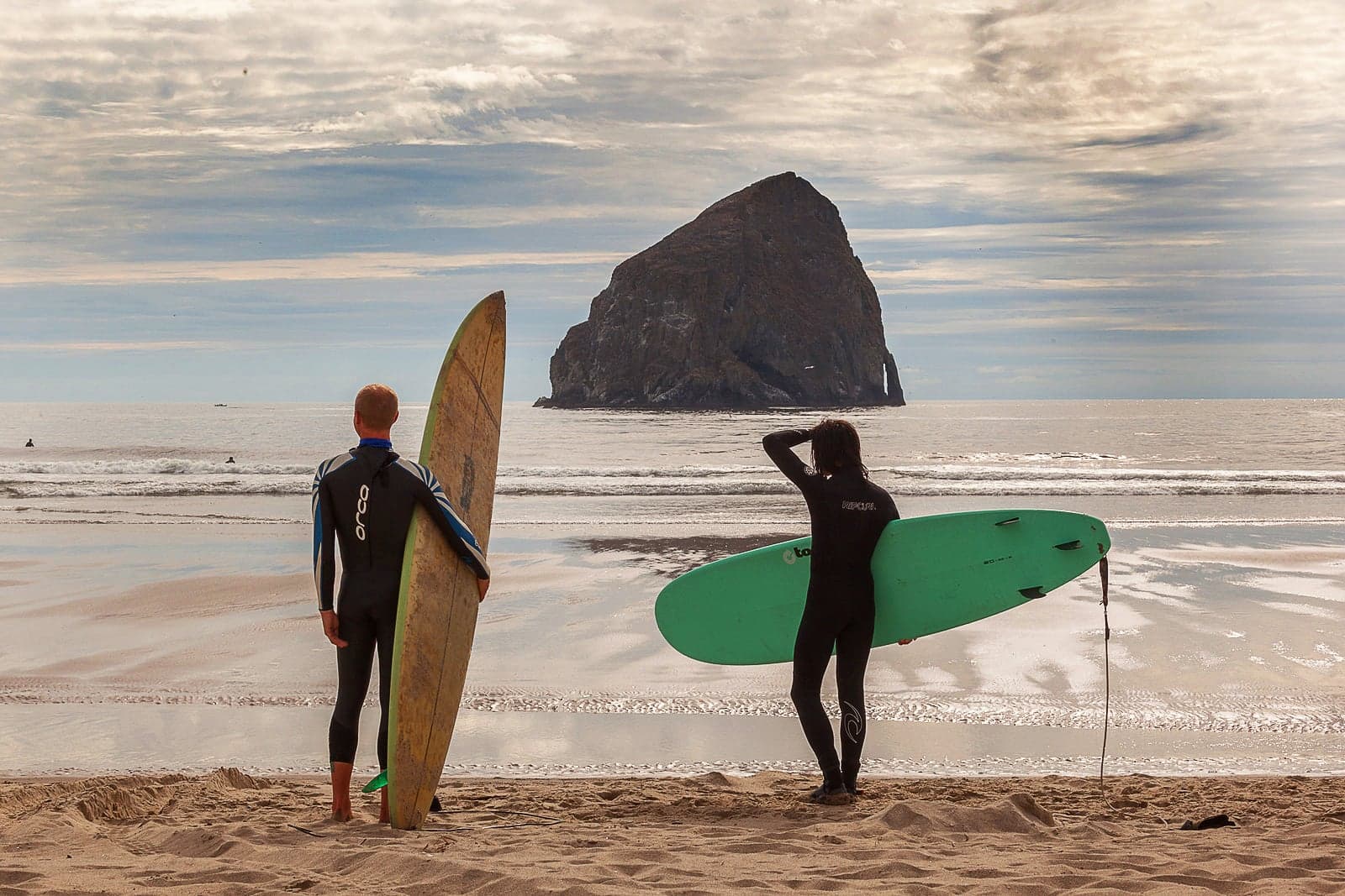 Two surfers with boards stand on a sandy beach, facing the ocean and a large rock formation under a cloudy sky, conveying a serene, adventurous vibe.