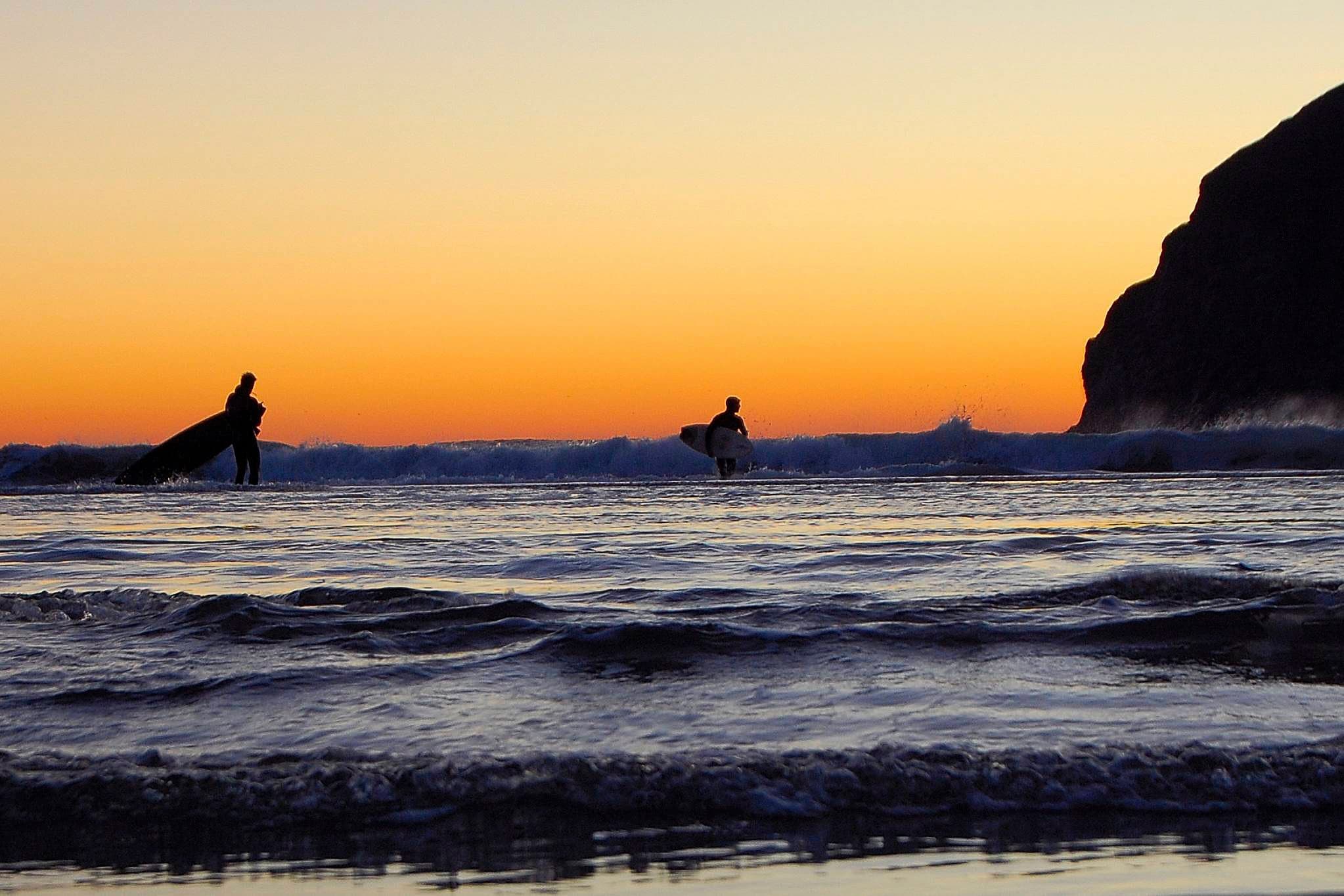 Two surfers carrying boards walk toward the ocean at sunset, creating silhouettes against a vibrant orange sky. Gentle waves and a rocky silhouette are visible.