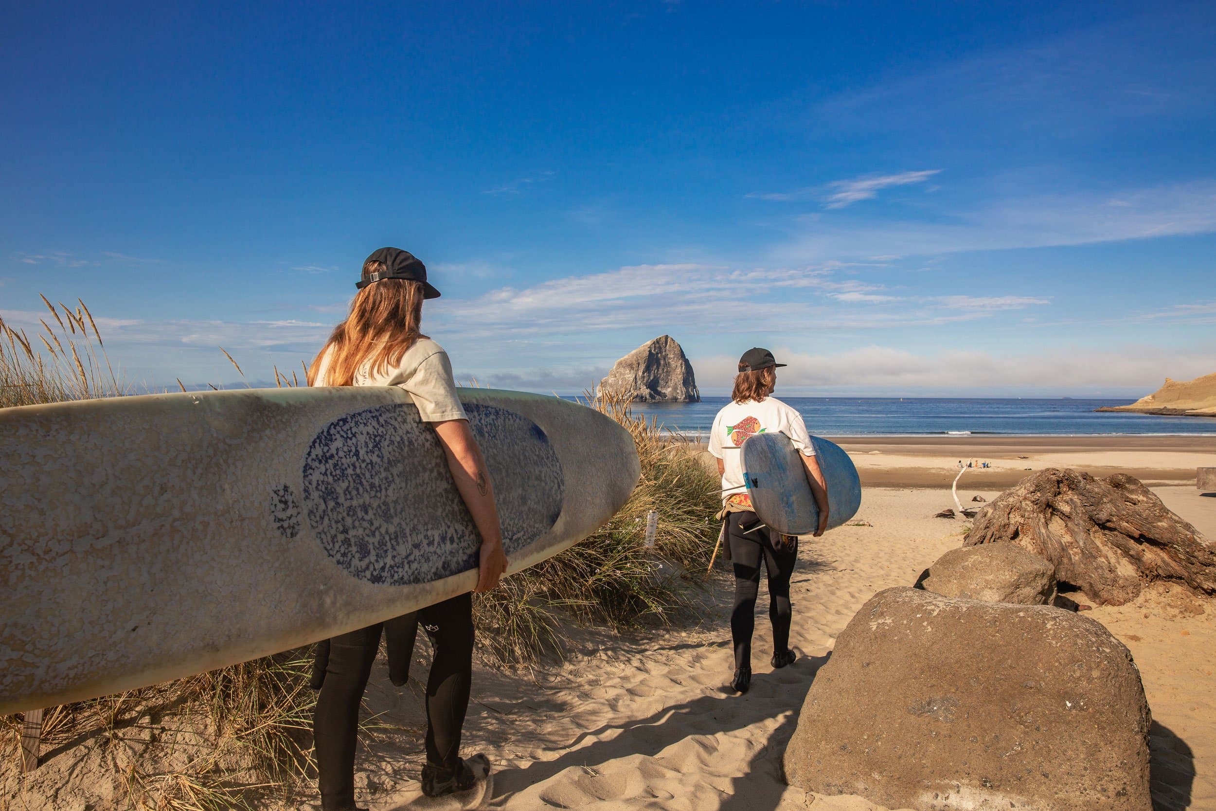 Two surfers holding surfboards walk toward the ocean on a sandy beach. The sky is clear, with a rocky island visible in the distance, conveying an adventurous and relaxed vibe.