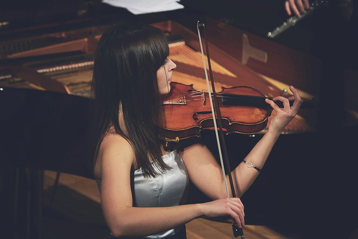 A woman in a white dress plays the violin passionately on stage. A piano and other musicians are in the background, creating an elegant atmosphere.