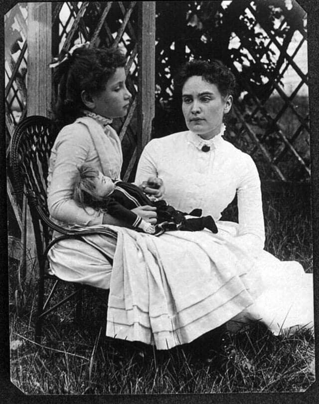 A girl holding a doll sits beside a woman on a wooden chair outdoors. Both wear long-sleeved Victorian dresses. The mood is calm, with lattice in the background.