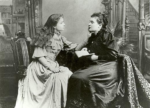 Two women sit closely, one in a dark dress holding a book. The other, in a light dress, leans in attentively. The Victorian setting feels calm and intimate.