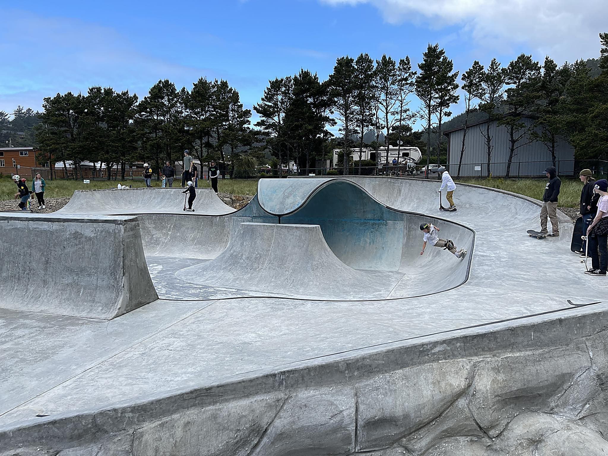Skaters and scooter riders carving the concrete bowls at the Pacific City skatepark on a bright day, a short ride from The Inn at Hart's Camp glamping resort on the Oregon Coast.